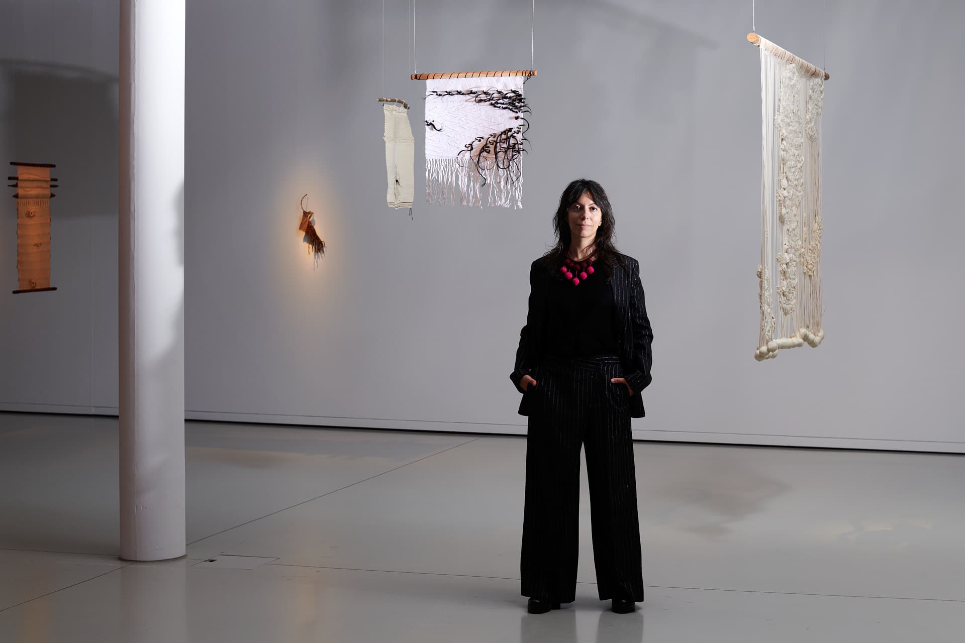 Portrait de la curatrice Elisa Rusca. Une femme habillée en noir se tient debout dans une salle d’exposition minimaliste, entourée de pièces textiles suspendues aux couleurs claires.