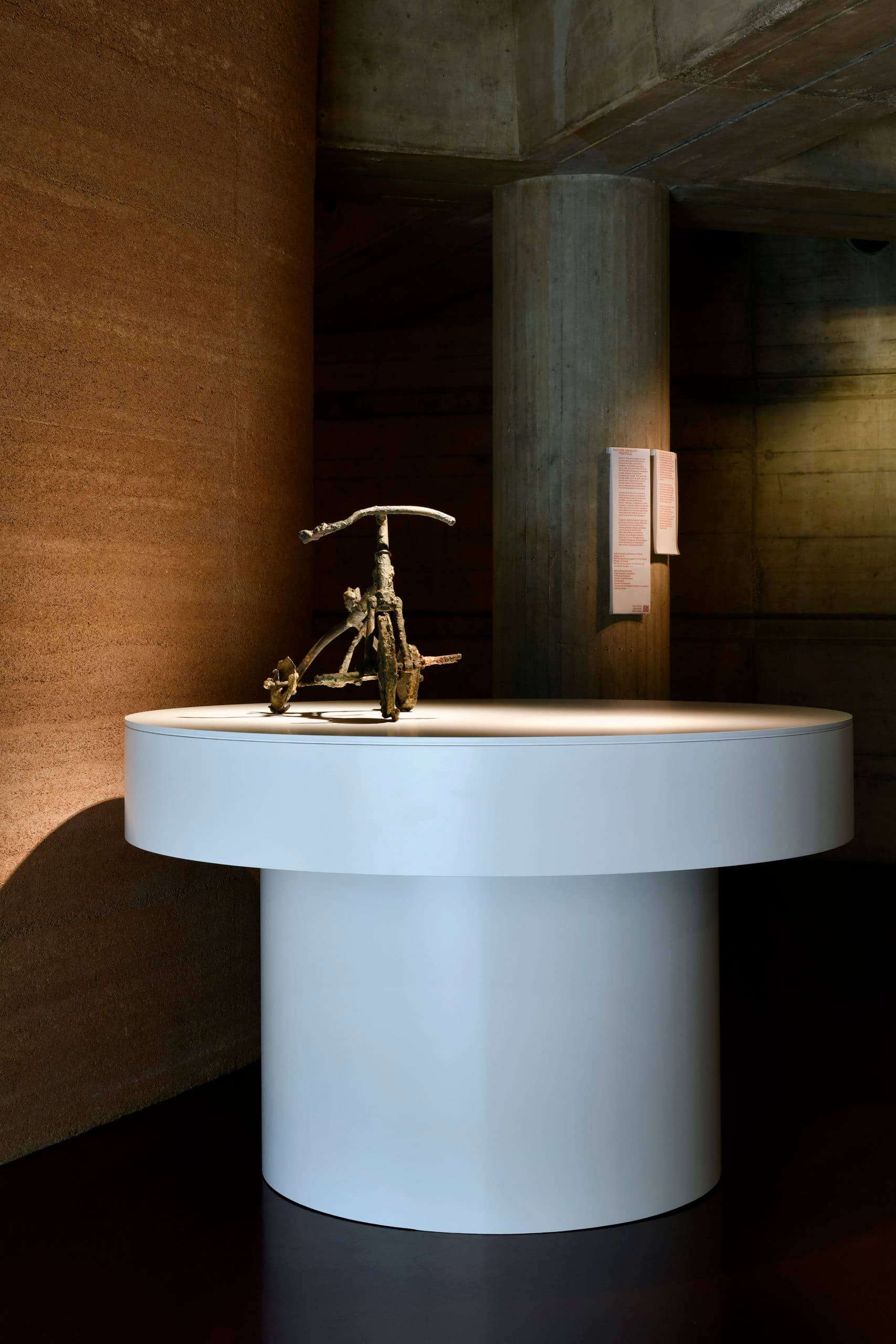 Sculpture d'un tricycle en métal exposé sur un large socle circulaire blanc mis en lumière par un projecteur. En arrière-plan, un mur texturé brun et une colonne de béton sur laquelle se trouve une signalétique explicative.