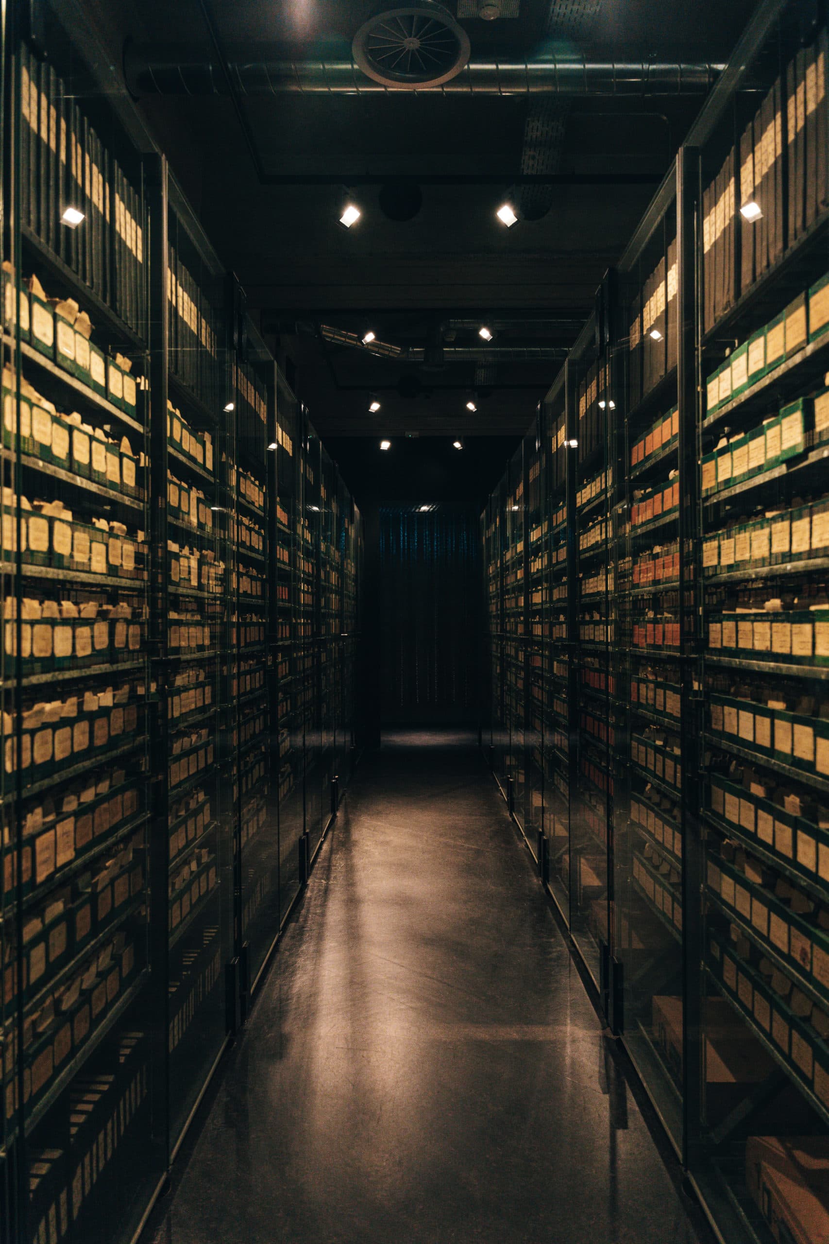 Couloir étroit bordé de hautes étagères vitrées remplies de boîtes d’archives étiquetées avec un éclairage tamisé.