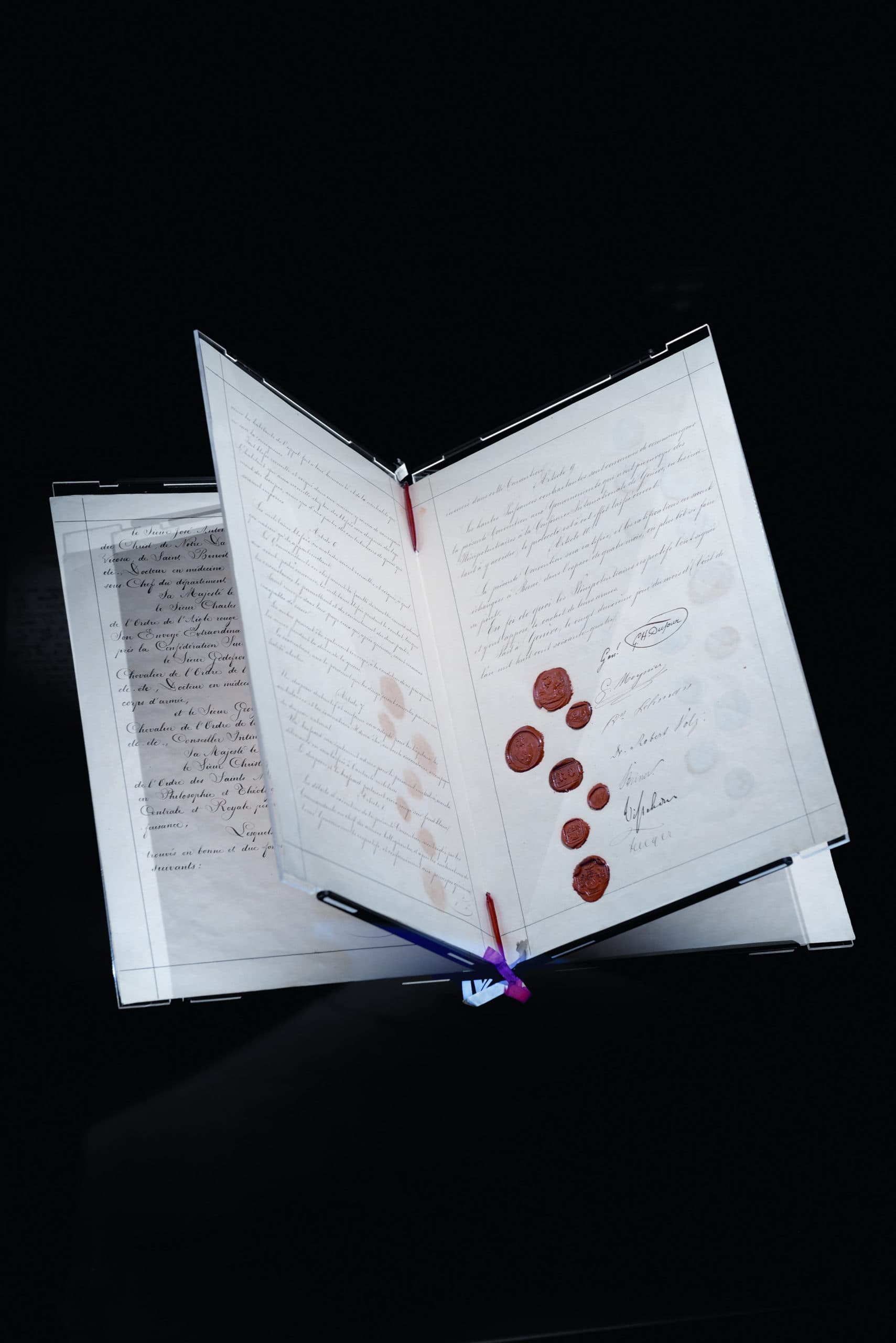 Documents manuscrits anciens ouverts en éventail, avec écriture cursive et sceaux de cire rouge, présentés sur un fond noir.