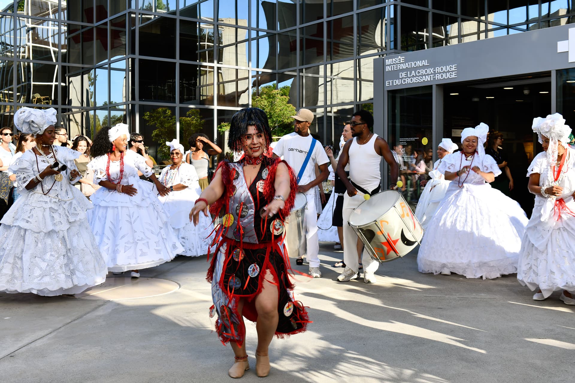 Une danseuse en costume rouge et noir mène une procession festive, entourée de percussionnistes et de personnes vêtues de robes blanches traditionnelles devant un bâtiment moderne.