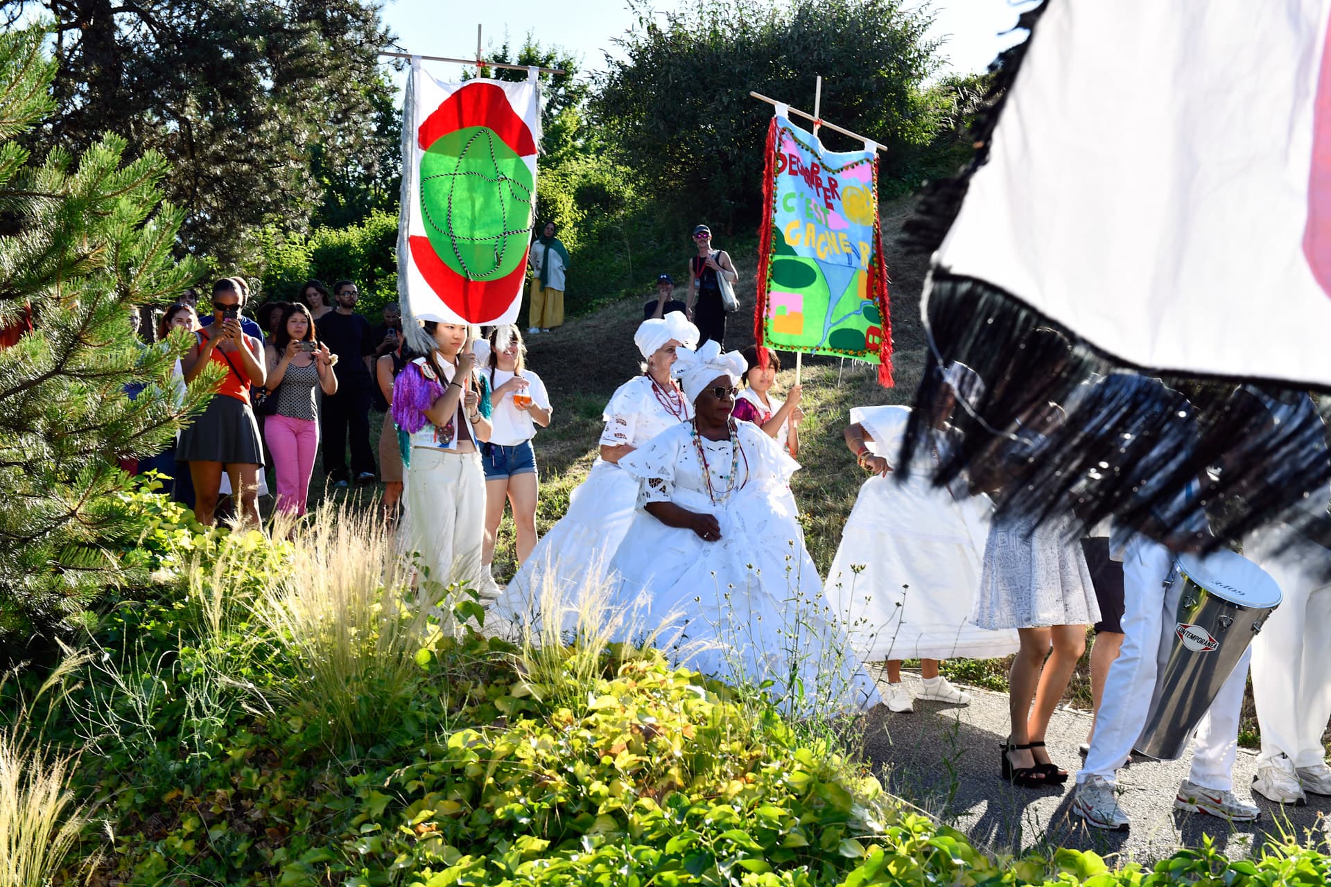 Une procession culturelle traverse un jardin avec des participants en robes blanches traditionnelles portant des bannières colorées et célébrant en plein air.