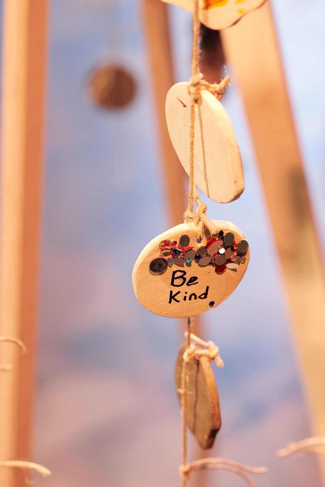 Amulette en céramique en forme de cercle suspendue à une ficelle avec d'autres cercles de céramique, décorées de strass de couleurs et portant l'inscription "Be Kind" en anglais.