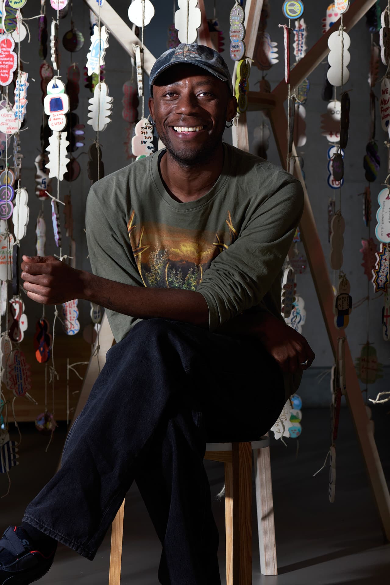 Un homme souriant assis devant une structure d'où pendent des amulettes en céramique attachées à des fils.
