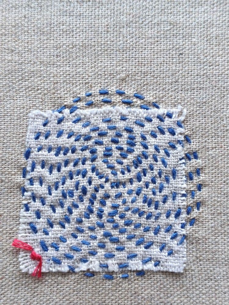 Points de broderies formant un cercle de fil bleu sur un tissus gris rapiécé avec un carré de tissus blanc.