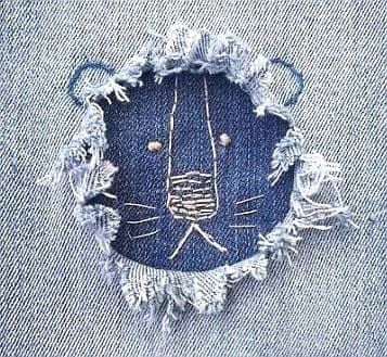 Gros plan sur un trou effiloché dans un jean clair, révélant un tissu bleu foncé avec une broderie décorative blanche dessinant un visage d'animal dans le trou.