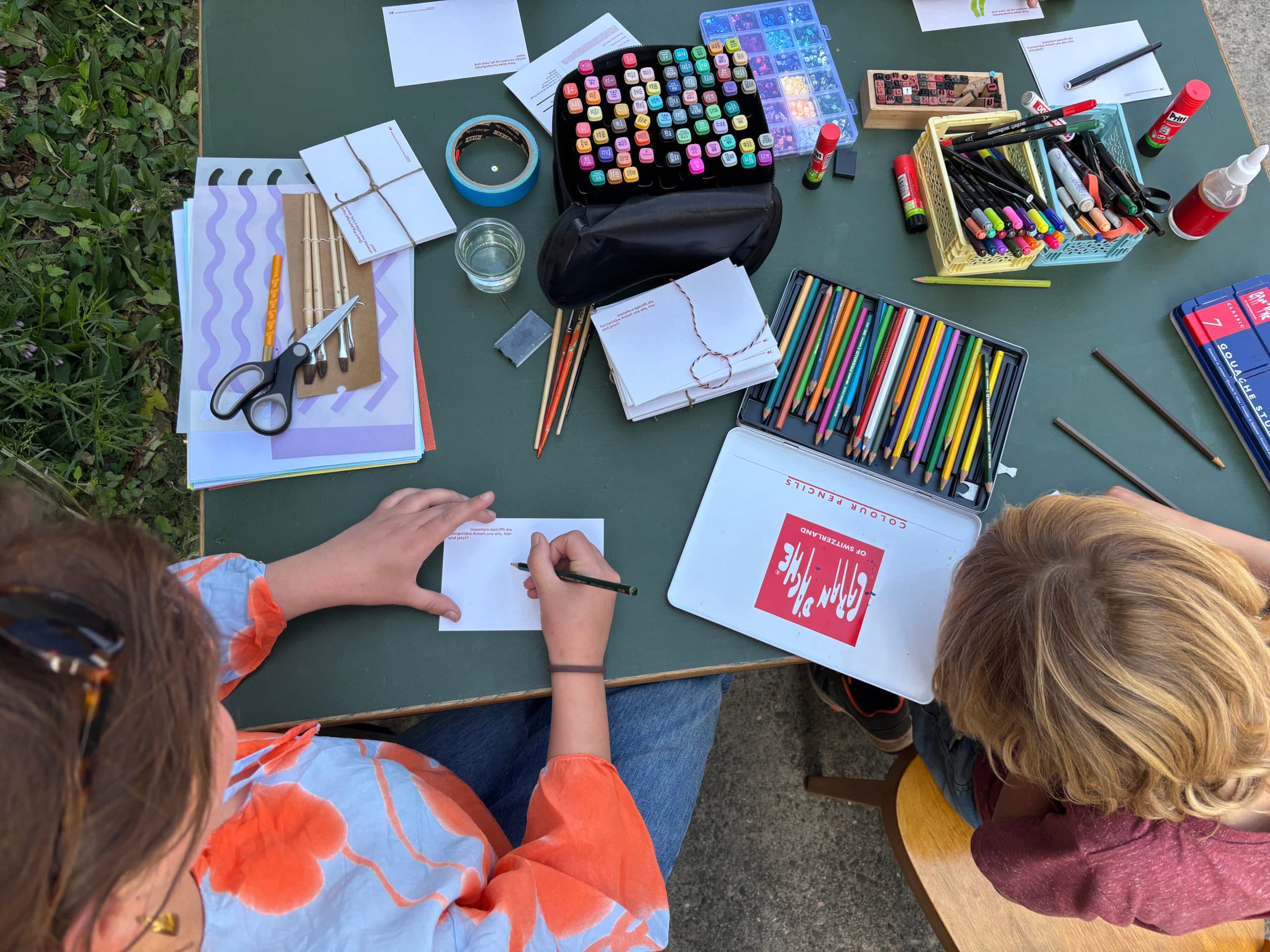 Une adulte et un enfant vus de haut en train de décorer des cartes postales vierges sur une table remplis de matériel de dessin et de bricolage tels que des crayons, de la colle et des feutres.