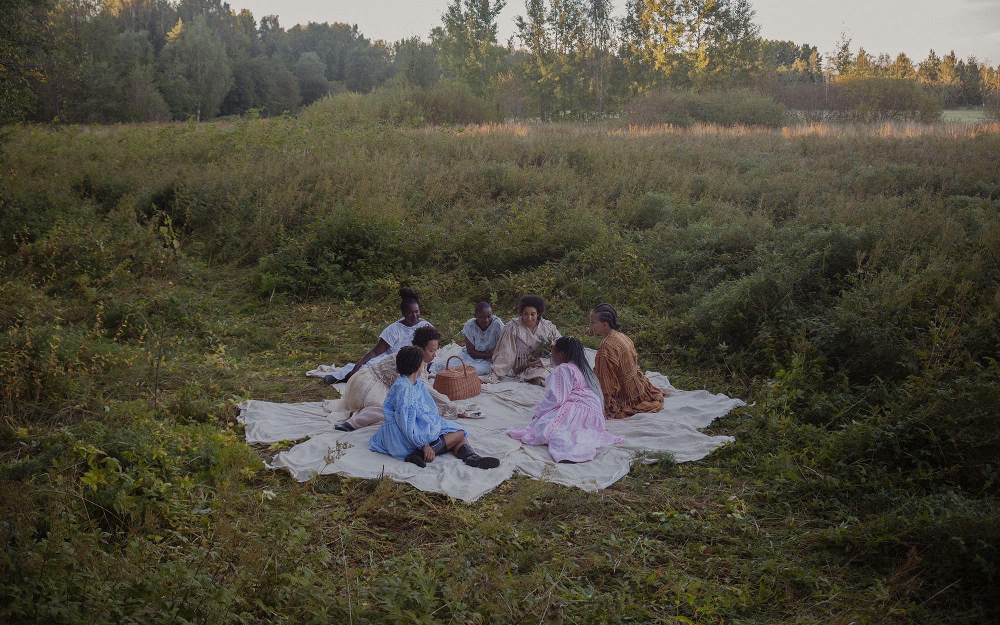 Femmes assises en cercle au milieu du champ de hautes herbes sur des draps blanc durant un picnic. L'atmosphère est douce et chaleureuse.