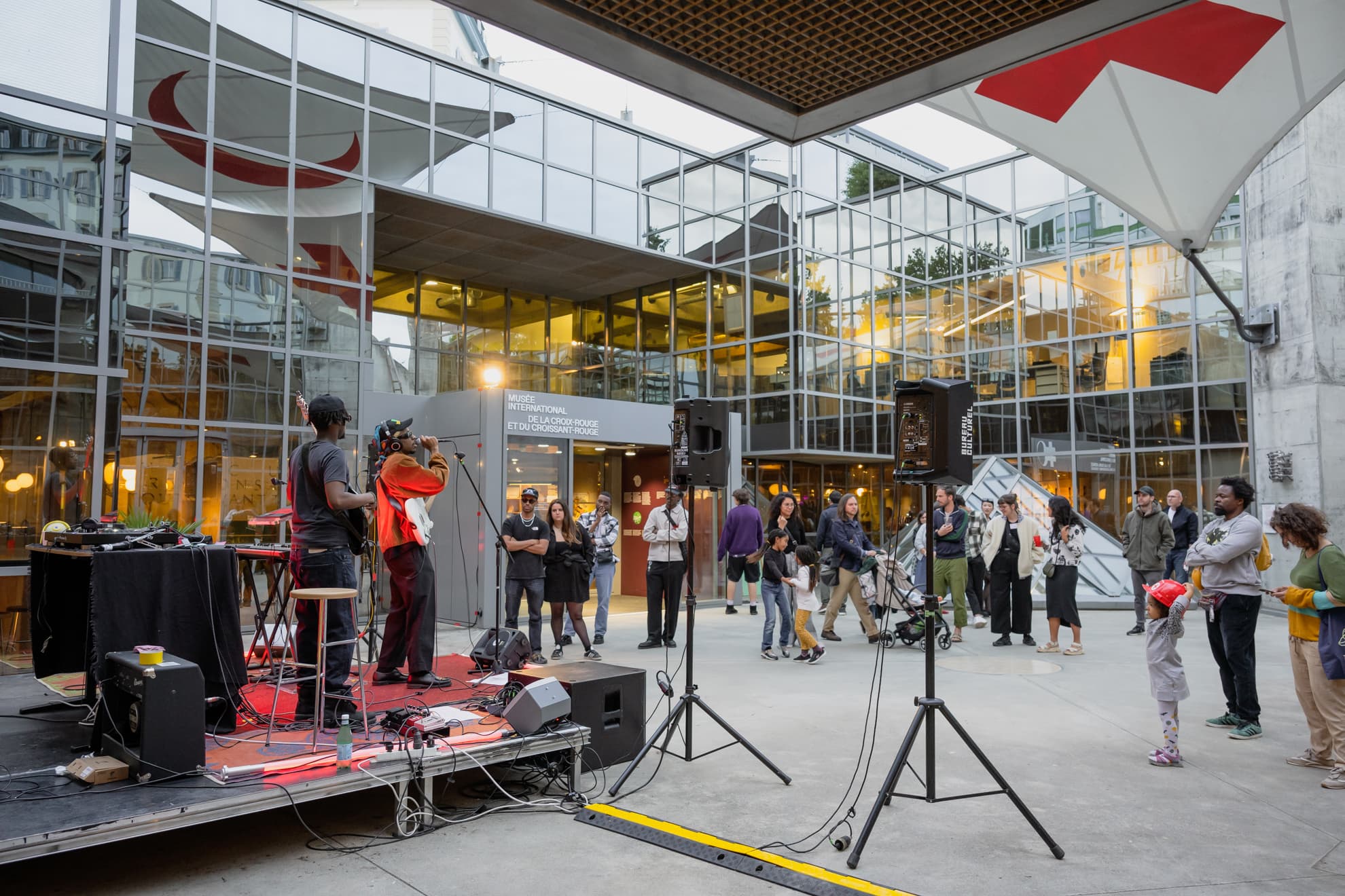 Personnes de tous âges écoutant un concert dans la cour intérieure d'un bâtiment vitré.