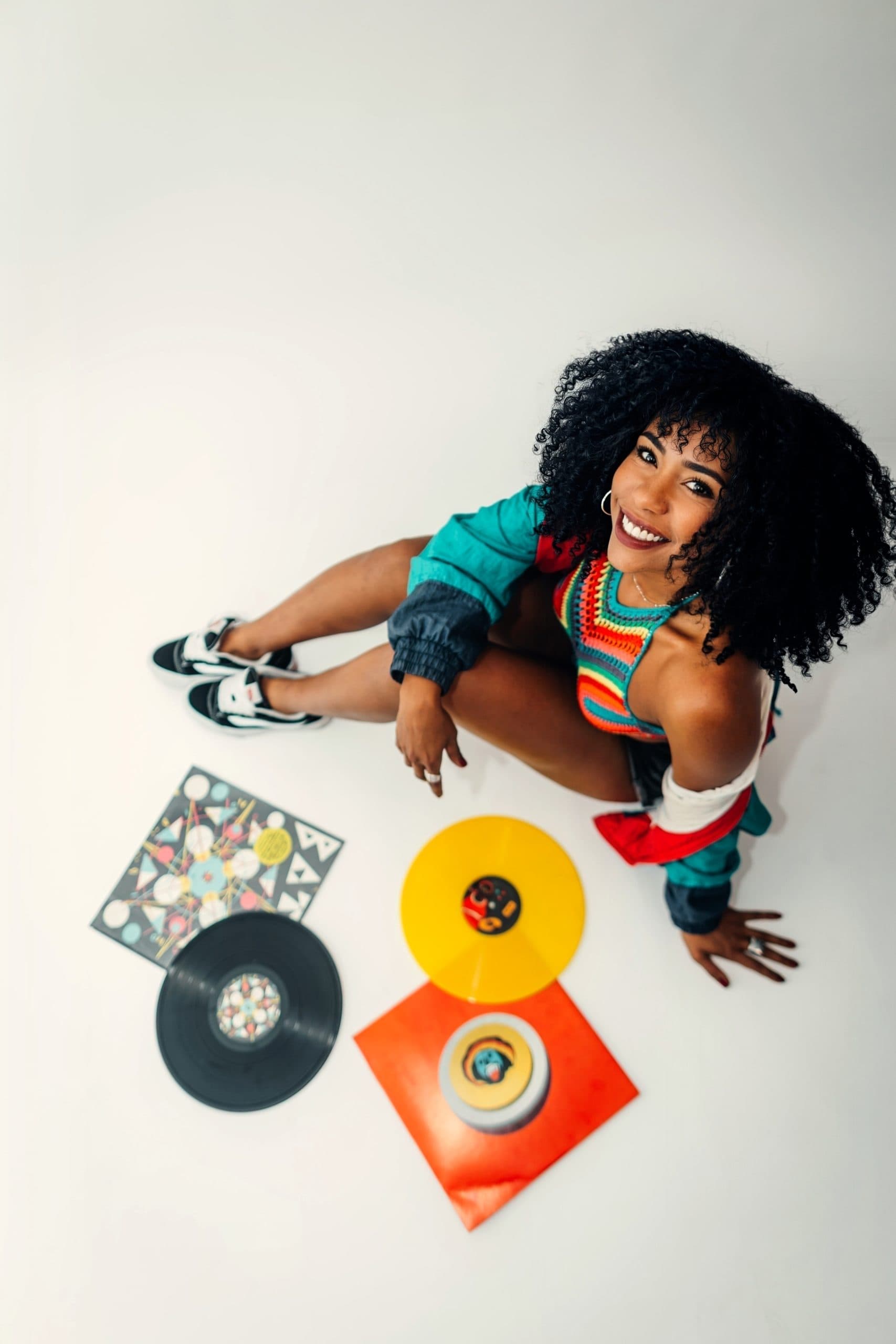 Une femme souriante et vêtue de vêtements colorés, prise en photo en plongé, assise sur un sol blanc entourée de vinyles et de pochettes de vinyles colorées.