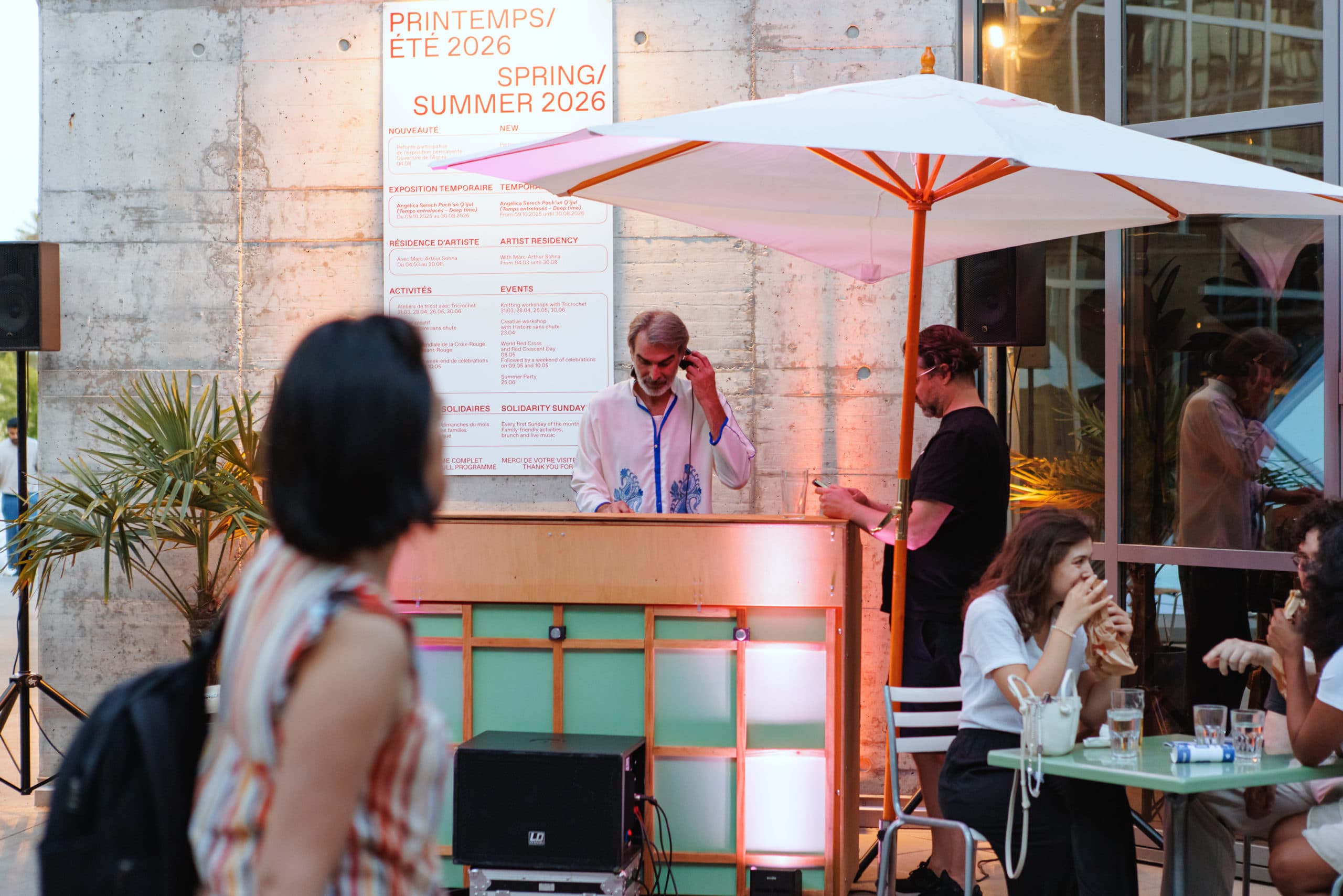 DJ derrière une console lors d’un événement en terrasse, sous un grand parasol blanc, tandis que des visiteurs discutent et prennent un verre autour de tables dans un espace contemporain éclairé par une lumière chaude.