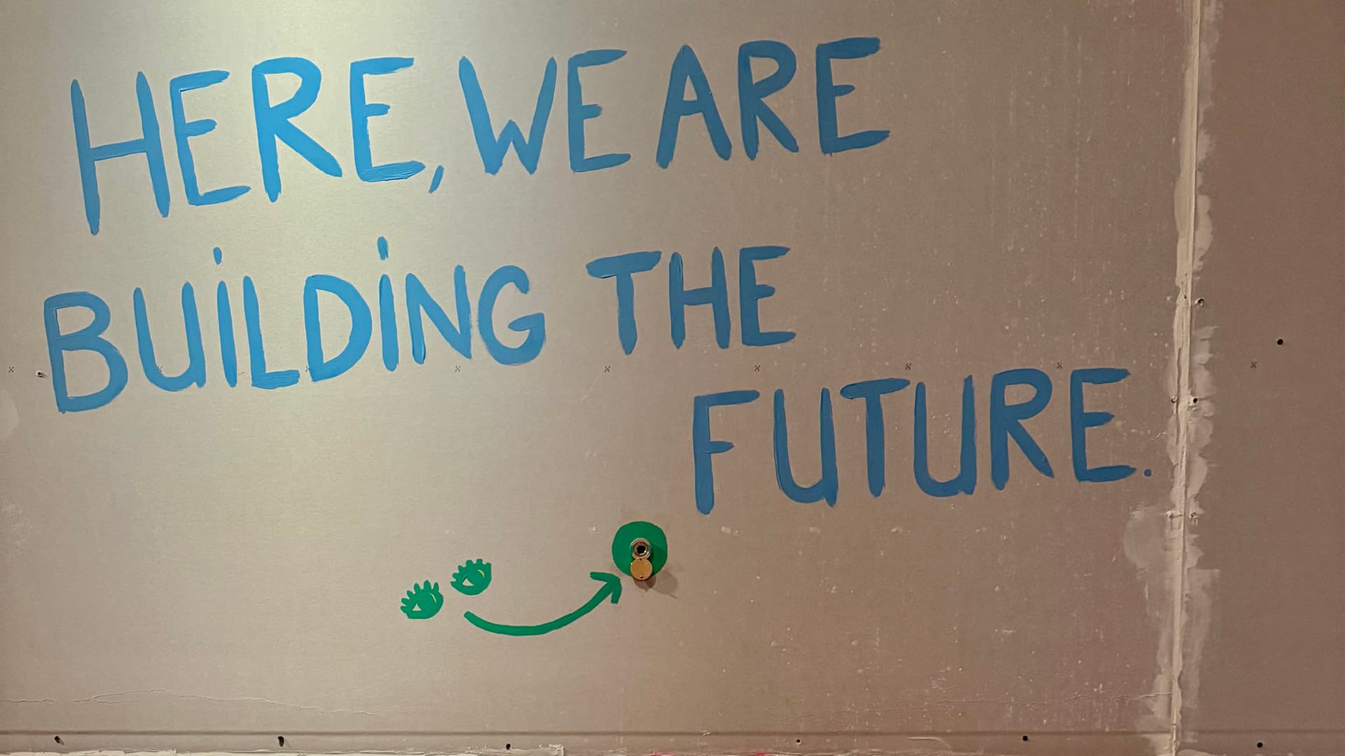 Texte en anglais "Here we are building the future" en bleu peint sur un mur gris, en dessous, deux yeux peints en vert et une flèche verte indiquent l'emplacement d'un judas.