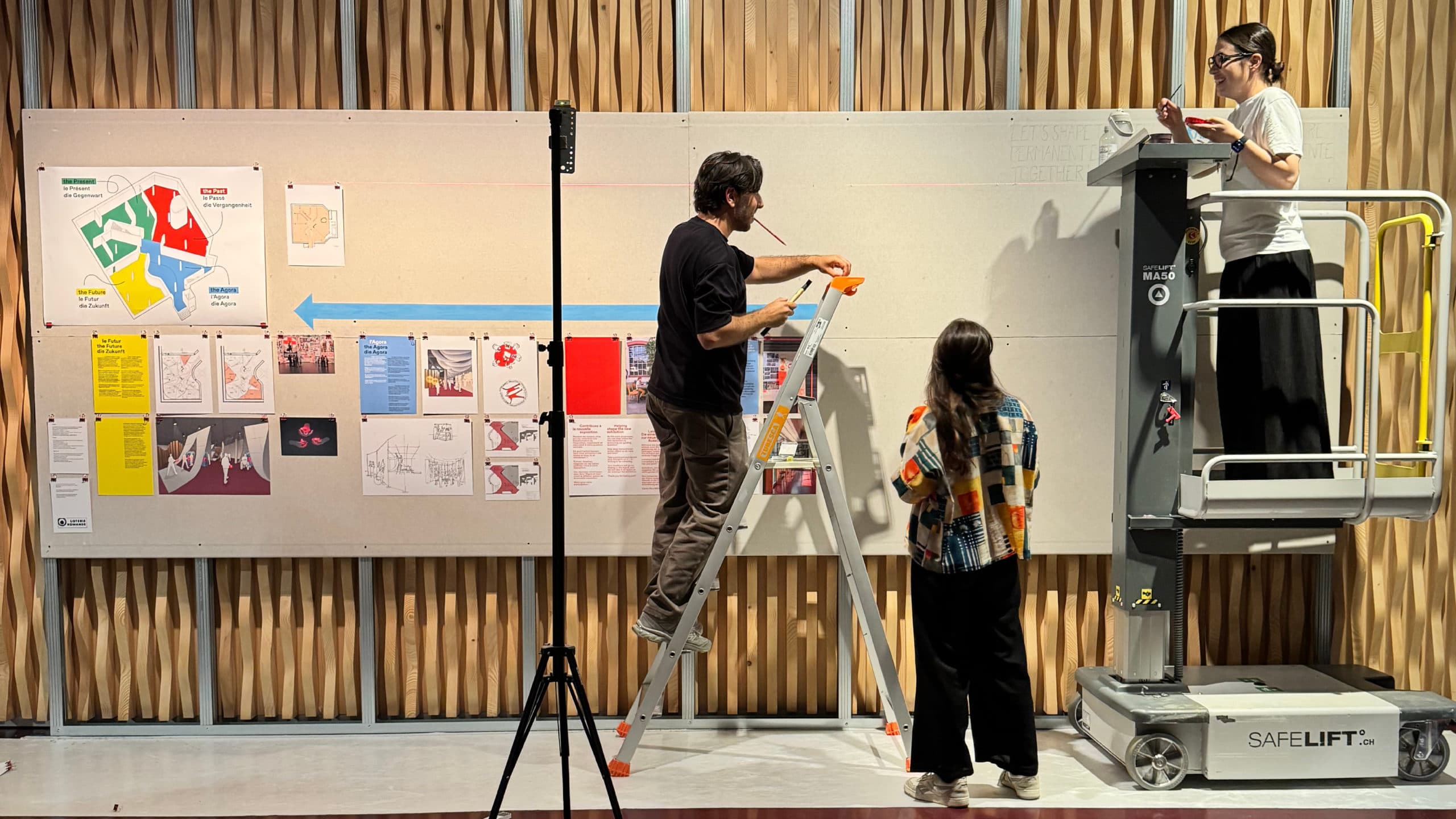 Trois personnes installent des affiches sur un grand panneau d’exposition intérieur : l’une est sur une échelle, une autre sur une plateforme élévatrice et une troisième se tient au sol. Le panneau présente des cartes, schémas et documents colorés, devant un mur à lattes de bois.